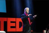 Michael Luongo speaking at TEDx Purdue Universit by Yash Trivedi Feb 18, 2023. Hands up, long hair, speaking with hands, velvet balck suit, red tie